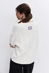 Sweater de punto color blanco off-white, con cuello redondo y diseño texturizado con trenzas y pompones en el frente.
