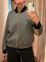 Chaqueta tipo bomber de lana con estampado de espiga en tonos grises. Presenta cuello, puños y ruedo en tejido elástico negro, cierre frontal dorado y bordado sutil en el pecho.