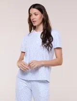 Pijama de dos piezas con estampado floral en tonos azules sobre fondo blanco, compuesto por una remera de manga corta con cuello redondo y botones frontales, y un pantalón capri con cintura elastizada y bolsillos.