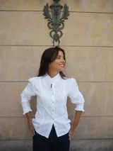 Camisa de manga larga confeccionada en broderie blanco, con diseño entallado, cuello alto y cierre frontal de botones. Presenta mangas abullonadas con hombros fruncidos y puños terminados en volados.