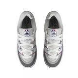 Zapatilla Adidas Adimatic de estilo skate, color gris claro con detalles en gris oscuro. Presenta tres franjas laterales en blanco, con ribetes finos en rojo y azul. La suela es gruesa y texturizada en la puntera.