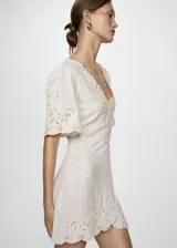 Vestido corto de color crudo con escote en pico y mangas cortas. Presenta un diseño de tejido con textura de cuadros y detalles bordados calados en el escote, las mangas y el ruedo. Cuenta con un corte recto y cierre de cremallera invisible en la parte posterior.