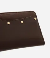 Billetera mediana color marrón, confeccionada en poliuretano con textura lisa y detalle de gamuza en la parte superior. Presenta cierre superior con cremallera dorada y aplicación de tachas metálicas decorativas en el frente.