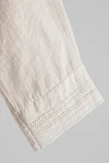 Camisa blanca de lino, con escote en V, mangas tres cuartos con detalles de encaje en los puños, cierre frontal de botones y diseño más largo en la parte trasera.