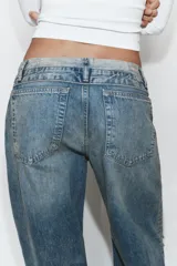Jeans de tiro medio con diseño de cintura doblada, cinco bolsillos y cierre frontal con cremallera y botón metálico. Presenta un acabado desgastado con roturas en las rodillas y corte recto.