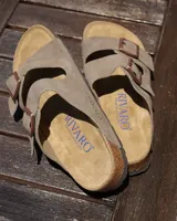 Sandalias tipo zueco marca Rivaro, confeccionadas en cuero gamuzado color beige. Presentan dos tiras anchas con hebilla metálica, plantilla anatómica de corcho y látex, y suela de goma antideslizante.