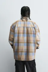Camisa de manga larga con cuello solapa y dos bolsillos frontales de plastrón con botón. Presenta un estampado de cuadros en tonos beige, azul claro y marrón.