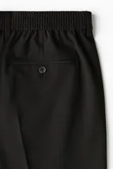 Pantalón de corte relajado color negro, con cintura elástica, cierre frontal con botón y cordón de ajuste oculto. Presenta piernas con raya marcada, bolsillos laterales al bies y bolsillos traseros ribeteados con botón.