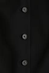 Chaleco largo negro, sin mangas, con cuello en V y cierre frontal de botones. Presenta bolsillos delanteros ribeteados y abertura en la parte trasera.