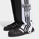 Pantalon deportivo Adidas modelo Adibreak de corte holgado, color negro con franjas laterales blancas y las icónicas tres tiras en negro. Presenta botones a presión a lo largo de los laterales y logo de la marca en la parte inferior.