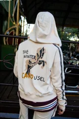 Campera con capucha de color crema, con diseño estampado en la espalda que incluye un caballo y la frase The Winter Carousel. Presenta bandas laterales con inscripciones en las mangas y un borde inferior con franjas horizontales en tonos bordó y celeste.