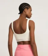 Top deportivo color blanco hueso, de corte ajustado, con breteles anchos y banda elástica inferior con inscripción. Presenta un pequeño recorte circular en el centro del busto.