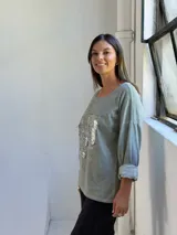 Remera de manga larga color verde, con cuello redondo y diseño holgado. Presenta un detalle frontal con parches de lentejuelas plateadas y estampado tipográfico.