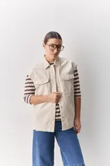 Chaleco de algodón color beige, de calce relajado, con cuello clásico, hombros amplios, cierre frontal con broches metálicos y bolsillos delanteros con solapa.