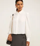 Camisa de crepé color blanco, con cuello inglés, manga larga abullonada con puños abotonados y cierre frontal oculto con botón diferenciado en el cuello.