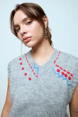 Chaleco de punto gris de corte holgado y largo cropped, con cuello redondo y sisas acanaladas. Presenta detalles bordados decorativos en la parte superior del pecho en tonos rojo y azul.