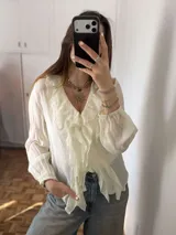 Blusa blanca de diseño ligero y fluido, con escote en V y volados que bordean la abertura frontal y el cuello. Cuenta con mangas largas abullonadas con puños elásticos y cierre delantero con botones.