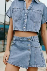 Camisa corta de denim en color azul claro, con cuello solapa, escote en V y manga corta. Presenta bolsillos delanteros de parche con solapa y cierre frontal de botones metálicos.