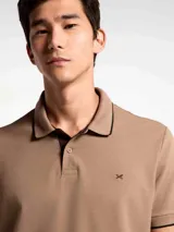 Chomba de piqué color tostado con cuello y puños en contraste negro. Presenta cierre frontal con botones y un pequeño bordado en el pecho.