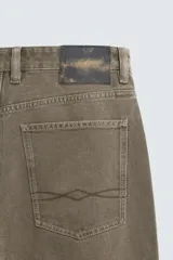 Pantalón de denim color verde oliva, estilo chino con corte ancho y bota acampanada (flare) desde la rodilla. Tiro medio, confeccionado en tejido rígido. Presenta bolsillos frontales y bolsillos traseros de plastrón.