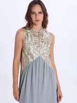Vestido largo sin mangas, de color negro con la parte superior bordada en tonos dorados y negros con patrones geométricos. Presenta escote en V y falda con volados escalonados.