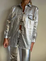 Camisa plateada brillante de manga larga con cuello clásico, cierre frontal con botones dorados y dos bolsillos con solapa en el pecho.