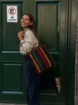 Cartera tipo tote de cuero con diseño de franjas verticales multicolores en tonos tierra, rojo y azul. Cuenta con correas trenzadas de cuero y cierre superior.