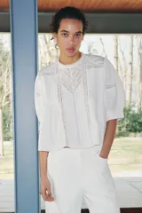 Blusa de algodón color blanco con cuello redondo y mangas abullonadas. Presenta detalles de puntilla a tono en el pecho y las mangas, bajo recto y cierre posterior con abertura y botón.