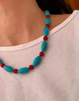 Collar de cuentas asimétrico compuesto por piezas alargadas de imitación piedra turquesa con vetas negras, intercaladas con pequeñas cuentas esféricas de color rojo. Incluye cadena extensora metálica en tono dorado.