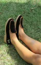 Mocasines de terciopelo verde oscuro, con ribetes en tono tostado y detalle de aplique metálico de un caballito en el talón. Poseen suela y taco bajo de madera.