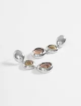 Caravanas colgantes de diseño moderno, compuestas por tres piezas circulares en degradado: una esfera metálica plateada superior, una piedra central en tono ámbar y una piedra inferior en tono ahumado, todas engarzadas en metal plateado.