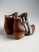 Botas cortas de cuero en tonos marrón y tostado, con paneles de cuero vacuno con estampado animal print tipo ponchada y detalles pintados a mano en dorado. Presentan punta fina, taco cuadrado de madera, costuras visibles a contratono y doble cierre lateral con tiradores metálicos personalizados.