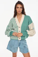 Cardigan tejido bicolor en tonos verde y crudo, con diseño de rayas verticales en la mitad derecha y liso en la izquierda. Presenta cuello en V, cierre frontal con botones y mangas abullonadas con puños ajustados.