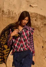Camisa de diseño original con combinación de telas escocesas en tonos rojo, azul y blanco. Presenta cuello camisero, cierre frontal con botones y volados decorativos en las sisas y en el dobladillo.