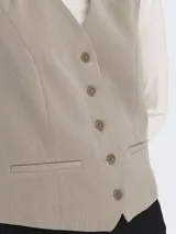 Chaleco sastrero color gris topo melange, de corte ajustado, con escote en V y cierre frontal de botones. Presenta bolsillos falsos en la parte delantera.