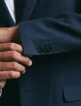 Saco de vestir azul marino con cuello solapa, manga larga y cierre frontal de dos botones. Presenta un diseño clásico con bolsillos laterales con solapa y un bolsillo superior en el pecho.