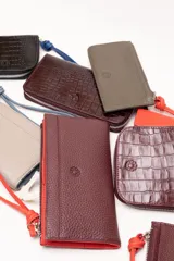Billetera rectangular de cuero con textura de reptil, cierre perimetral y correa de muñeca lateral. Presenta un pequeño aplique metálico circular en el centro.