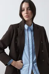 Camisa blanca de manga larga con detalles bordados en contraste a lo largo de la botonadura, el cuello y los puños. Presenta una silueta relajada y un diseño clásico con un toque artesanal.