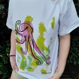 Remera blanca con estampado artístico de pulpo en tonos rosados y salpicaduras verdes, de corte holgado y mangas cortas.