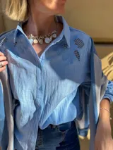 Camisa blanca de corte amplio con mangas tres cuartos abullonadas. Presenta detalles de bordado calado en los hombros y en las mangas, con cierre frontal de botones.