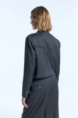 Blazer cropped de sarga con diseño a rayas verticales, corte entallado y cierre frontal cruzado con botones. Presenta detalles desflecados en los hombros y en el canesú trasero, además de flecos de seda desmontables en la solapa.