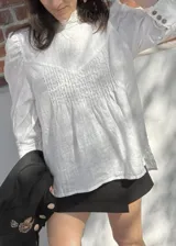 Blusa de lino color blanco con cuello alto, detalle de alforzas en el frente y mangas abullonadas. Presenta cierre posterior con botones y puños con detalle de puntilla negra.
