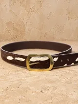 Cinturón ancho de cuero con textura de pelo y estampado animal print de manchas marrones y blancas. Cuenta con hebilla rectangular metálica con terminación envejecida y perforaciones regulables.