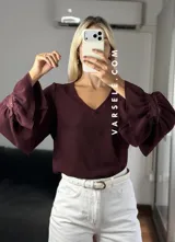 Blusa de tela cey color bordó, con escote en V y mangas largas abullonadas que terminan en puños con detalle de encaje y volados.