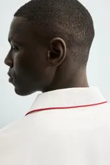 Pijama de dos piezas confeccionado en popelín de algodón blanco, con vivos de ribete en color bordó. Incluye una camisa de manga larga con cuello bowling, bolsillos de plastrón y cierre frontal de botones, y un pantalón de corte relajado con cintura elástica y bolsillos laterales.