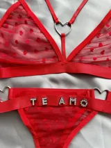 Conjunto de lencería rojo de tul con diseño de corazones aterciopelados en relieve. Incluye un soutien tipo triángulo con argollas metálicas en forma de corazón y tiras regulables, y una bombacha colaless con tiras laterales ajustables unidas por argollas en forma de corazón y el texto "TE AMO" aplicado con apliques de pedrería brillante.