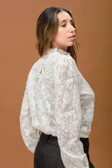 Camisa de encaje color crudo, con cuello alto con volados, mangas largas abullonadas y puños elásticos.