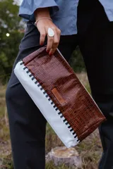 Sobre tipo baguette de cuero con textura croco en color marrón. Presenta un diseño bicolor con un lateral en cuero blanco unido mediante un trenzado artesanal en color negro. Incluye una etiqueta de cuero con el logo de la marca y un lazo desmontable.