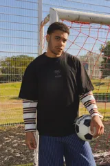 Remera negra de corte oversize con estampa frontal en color gris desgastado que hace referencia a la historia de la selección uruguaya. Presenta mangas largas integradas con diseño de rayas horizontales en tonos blanco, marrón y negro.