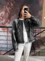 Campera de cuero genuino color negro, con diseño de cuello redondo y cierre frontal con botones a presión. Presenta bordados decorativos en gamuza con formas geométricas en el frente y detalles de costuras en forma de cruz en el ruedo y los puños.